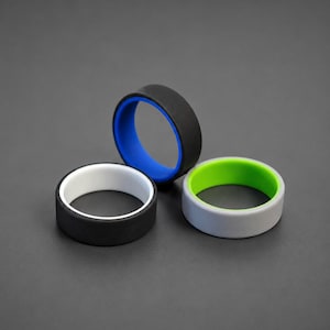 May include: Three silicone rings arranged in a cluster. One ring is black with a white interior, one is black with a blue interior, and one is light grey with a bright green interior. Each band features a smooth, matte finish and a flat profile.