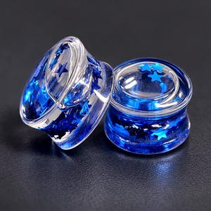 May include: Two clear, acrylic ear gauges filled with blue star-shaped glitter. The gauges are cylindrical with a flared edge, and the glitter is suspended within the clear material. The background is a dark, solid color.