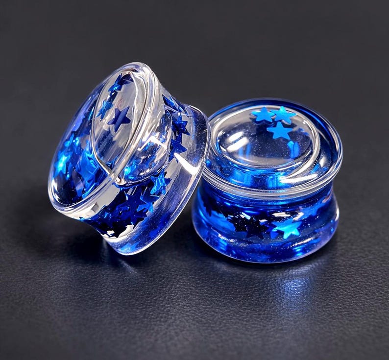 May include: Two clear, acrylic ear gauges filled with blue star-shaped glitter. The gauges are cylindrical with a flared edge, and the glitter is suspended within the clear material. The background is a dark, solid color.