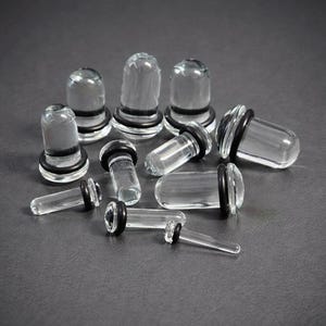 May include: A selection of clear glass ear plugs with black rubber O-rings. The plugs are of various shapes, some with rounded ends and others cylindrical. The plugs are displayed on a dark grey surface.