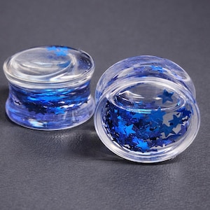 May include: Two clear, glass ear gauges with blue star-shaped glitter inside. The gauges have a rounded shape and are designed to be worn in stretched ear piercings. The glitter creates a sparkling effect.