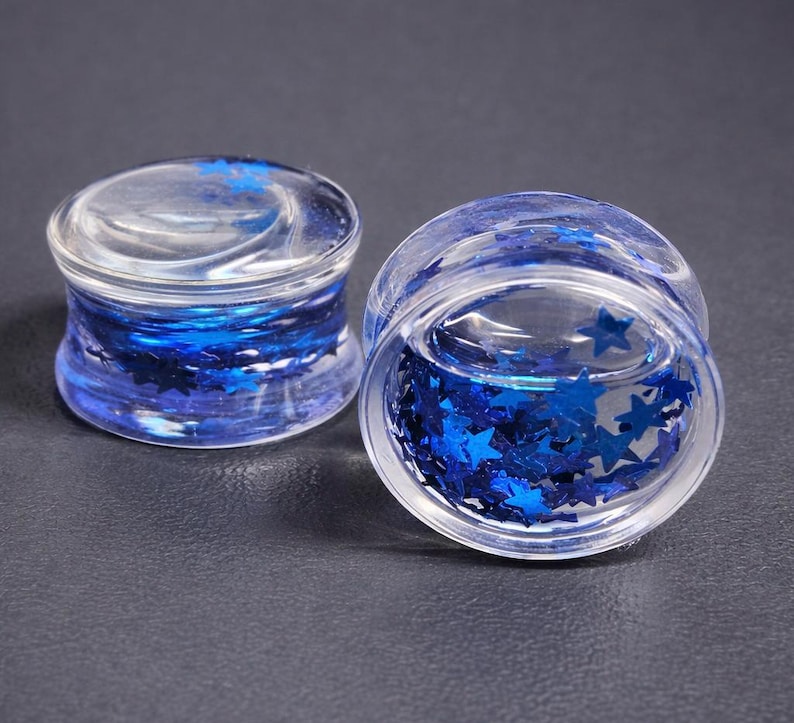 May include: Two clear, glass ear gauges with blue star-shaped glitter inside. The gauges have a rounded shape and are designed to be worn in stretched ear piercings. The glitter creates a sparkling effect.