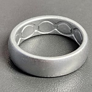 May include: A silver-colored ring with a matte finish. The inside of the ring features a series of interconnected oval shapes. The ring is a simple band design, suitable for everyday wear.