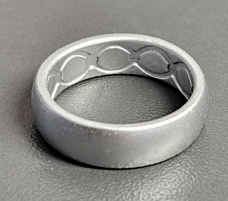 May include: A silver-colored ring with a matte finish. The inside of the ring features a series of interconnected oval shapes. The ring is a simple band design, suitable for everyday wear.
