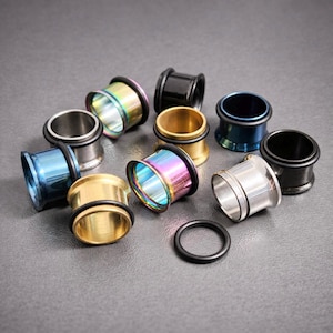 May include: An assortment of ear gauges in diverse colours, such as silver, gold, black, and iridescent rainbow. Each gauge features a flared design with a black rubber O-ring. The gauges are arranged on a grey surface.