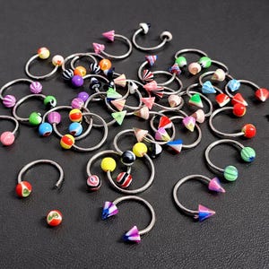 Può includere: Una collezione di gioielli per il corpo a forma di ferro di cavallo argentati con elementi decorativi colorati. L'assortimento include pezzi con perline vibranti, palline a righe e accenti a forma di cono in vari colori.