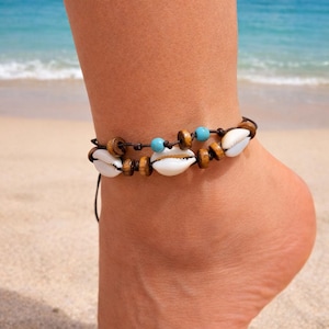 Könnte beinhalten: Fußkettchen mit weißen Muscheln, Holzperlen und türkisfarbenen Perlen an einer dunkelbraunen Kordel. Der Schmuck wird am Knöchel getragen, mit Strand und Meer im Hintergrund.