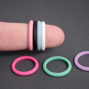 May include: A selection of colourful silicone rings in pink, black, light blue, white, mint green, and lavender. Some rings are stacked on a finger, while others are displayed separately on a dark grey surface. These rings are likely used as accessories.