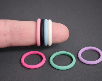 May include: A collection of colorful silicone rings in pink, black, light blue, white, mint green, and lavender. Some rings are stacked on a finger, while others are displayed separately on a dark gray surface. These rings are likely used as accessories or for other purposes.