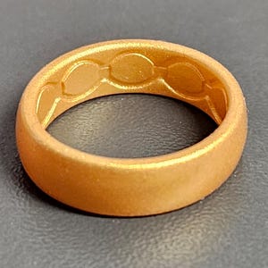 May include: A gold-colored ring with a textured surface. The inside of the ring features a series of three connected oval shapes. The ring is made of a flexible material and has a matte finish.