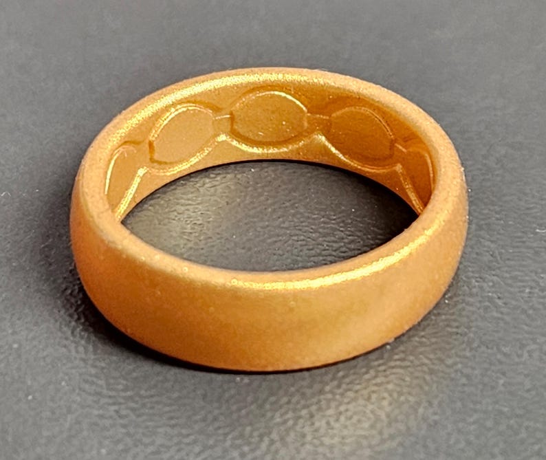 May include: A gold-colored ring with a textured surface. The inside of the ring features a series of three connected oval shapes. The ring is made of a flexible material and has a matte finish.