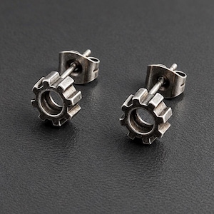 May include: A pair of stud earrings featuring a dark silver-toned gear or cogwheel design. Each earring has a circular center with radiating teeth and includes a standard post and butterfly clutch backing for secure attachment.