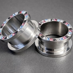 Può includere: Due dilatatori per orecchie color argento con un anello di strass colorati. Gli strass sono rosa, blu e neri. I dilatatori sono cilindrici con un bordo svasato.