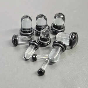 Könnte beinhalten: Eine Sammlung klarer Glas-Ohrstöpsel mit schwarzen Rändern und Basen. Die Stöpsel haben an einem Ende eine abgerundete, kuppelförmige Form und am anderen Ende eine zylindrische Form. Die Stöpsel sind auf einer grauen Oberfläche angeordnet.