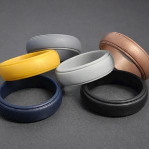 May include: A selection of silicone rings in shades of yellow, navy blue, grey, black, and bronze. The rings are displayed on a dark grey surface, highlighting their smooth, rounded design. The image showcases a variety of colours.
