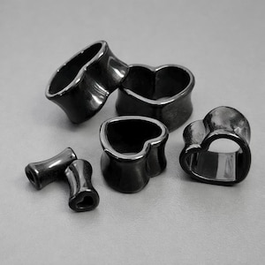 Peut inclure: Plusieurs écarteurs d'oreille noirs en forme de cœur. Les écarteurs sont fabriqués dans un matériau sombre et brillant et sont de tailles différentes. La conception présente une forme de cœur avec un bord évasé, adaptée aux piercings d'oreille étirés.