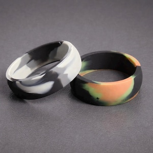 May include: Two silicone rings. One ring has a black, white, and grey camouflage pattern. The other ring has a camouflage pattern in black, orange, and green. Both rings are on a grey surface.