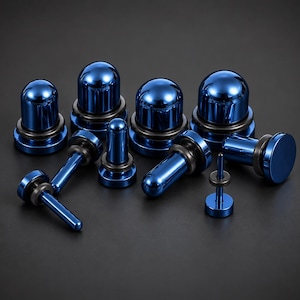 May include: A collection of metallic blue body jewellery pieces, including several rounded plugs and elongated barbell-style studs. Each piece features a smooth, reflective finish and is accented with a thin black rubber O-ring for secure placement.