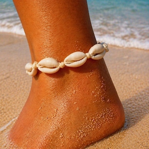 Peut inclure: Une chevillère composée d'une série de coquillages cauris blanc cassé enfilés sur un cordon de couleur claire. Les coquillages sont disposés en une rangée continue, mettant en valeur leurs formes ovales naturelles, lisses et allongées.