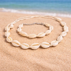 Könnte beinhalten: Ein cremefarbenes Muschelhalsketten- und Armband-Set. Die Halskette und das Armband bestehen aus kleinen, ovalen Muscheln, die mit einer cremefarbenen Kordel aufgereiht sind. Das Armband hat einen silberfarbenen Kettenverschluss. Das Set wird an einem Sandstrand präsentiert.