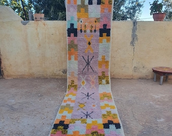 May include: A long, rectangular rug with a colorful geometric pattern. The rug is made of wool and has a fringe on the ends. The colors in the rug include pink, orange, yellow, green, and black.