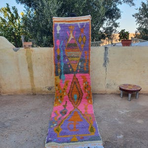 Puede incluir: Una alfombra larga y colorida con un patrón geométrico en tonos de rosa, morado, naranja y amarillo. La alfombra tiene flecos en el borde inferior.