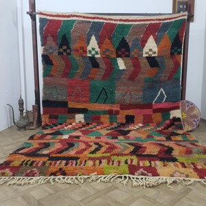 May include: A vibrant, hand-woven rug with a geometric design. The rug features a mix of colors including red, green, orange, and black. The rug has a fringed edge and is displayed on a wooden frame. A decorative hat is next to the rug.