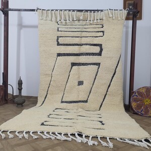 May include: Cream-colored, geometric patterned rug with black lines and a fringe. The rug is displayed on a metal rack. A decorative pillow with a floral design is visible in the background.