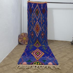 Puede incluir: Una alfombra larga y azul vibrante con un patrón geométrico de diamantes en tonos de rojo, naranja, verde y rosa. La alfombra tiene un borde con flecos y se muestra verticalmente, mostrando su intrincado diseño y textura.