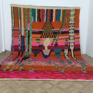 May include: A vibrant, hand-woven rug with a colorful geometric design. The rug features a mix of orange, pink, green, and blue hues. The rug's texture is visible, with a thick pile and a variety of patterns. This decorative rug is suitable for any room.