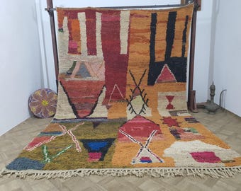 May include: A large, colorful rug with a geometric design. The rug features a variety of shapes and colors, including red, orange, black, and white. The rug has a fringe along the bottom edge and is displayed on a wooden rack.