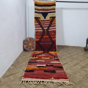 May include: A long, rectangular, hand-woven rug with a geometric design. The rug features a vibrant color palette of red, orange, yellow, and black. The design includes diamond and stripe patterns. The rug has a fringed edge and is displayed on a wooden floor.
