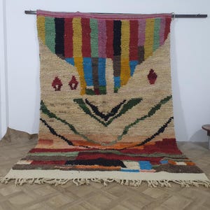 May include: A hand-woven, rectangular rug with a beige background and a colorful geometric design. The top section features vertical stripes in red, yellow, green, and blue. The bottom has a mix of colors, including red, orange, and black. The rug has a fringe along the bottom edge.