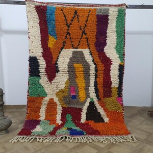 May include: A vibrantly colored, rectangular rug with a textured, woven design. The rug features a mix of orange, white, green, and purple hues, with a geometric pattern in the center. The rug has a fringe along the bottom edge.