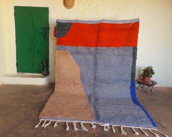 May include: A hand-woven rug with a geometric design in shades of blue, orange, brown, and gray. The rug has a fringe border.
