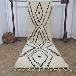 May include: Cream-colored, rectangular rug with a geometric black diamond pattern. The rug has a textured, woven appearance and a fringe along the bottom edge. The design features a series of connected diamond shapes.
