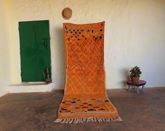 May include: A long, rectangular, orange rug with a geometric pattern in shades of blue, red, and yellow. The rug has a fringe border and is laid out on a concrete floor.