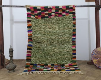 May include: A rectangular, hand-woven rug with a central field of light green. The rug features a colorful border with a checkerboard pattern in shades of pink, orange, and brown. The rug has a fringe along the bottom edge.