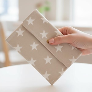 Op de afbeelding: Een beige stoffen portemonnee met een wit sterrenpatroon. De rechthoekige portemonnee heeft een klep die sluit met een goudkleurige drukknop. De portemonnee wordt door een hand vastgehouden.