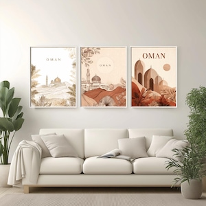 May include: Three framed art prints featuring watercolor illustrations of Oman. Each print has a white border and depicts a different landscape scene with the word "OMAN" at the top. The color palette includes shades of brown, beige, and green.