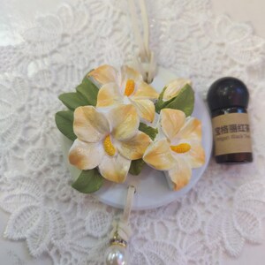 Könnte beinhalten: Runder, weißer Keramikschmuck mit drei gelb-orangenen Hibiskusblüten und grünen Blättern. Eine kleine, dunkle Flasche mit Text befindet sich neben dem Schmuck. Ein cremefarbenes Band und eine Perle hängen an dem Schmuck.