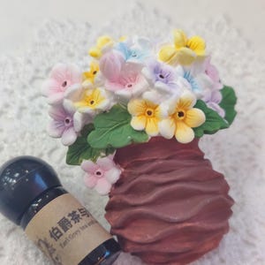 May include: A brown ceramic vase with a bouquet of pastel-colored flowers. The flowers are pink, yellow, and blue. A small black bottle with text is next to the vase. The vase is likely made of ceramic.
