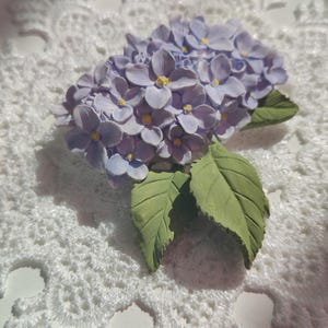Puede incluir: Broche artesanal de flor de hortensia lavanda con centros amarillos y hojas verdes. El diseño floral se encuentra sobre un fondo blanco similar al encaje, creando una estética delicada y artística. Este broche es una obra de arte portátil.
