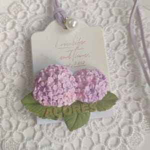 以下が含まれることがあります： ラベンダー色の紐とパールのアクセントが付いた、タグ型のペンダント。ペンダントには、ピンク色の紫陽花の2つの花、緑の葉、そして「ACORES」の文字が特徴です。タグには「I love life and time.」と書かれています。