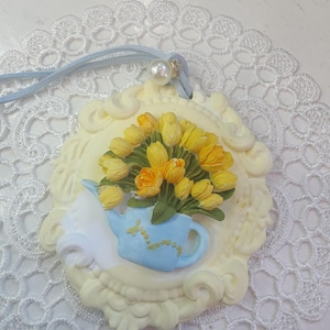 May include: A cream-colored pendant with a floral design. The pendant features a blue teapot filled with yellow tulips and a cream-colored frame. A light blue cord and a pearl bead are attached to the pendant.