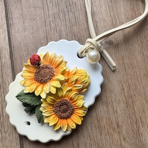 May include: A white, scalloped-edge tag featuring a 3D sunflower design with yellow and orange petals, brown centers, and green leaves. A small red ladybug rests on one flower. A cream-colored cord with a pearl accent is attached.