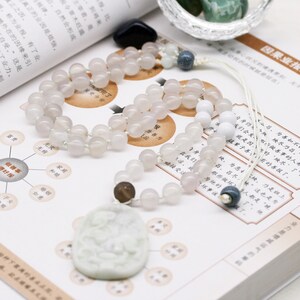 May include: A white jade pendant with a carved design hangs from a white and light pink beaded necklace. The necklace is on a book with Chinese text. There are also colorful stones in a glass bowl.