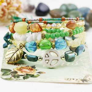 May include: A set of six colorful beaded bracelets with a peace sign charm. The bracelets feature green, blue, white, and yellow beads, and a gold leaf charm.