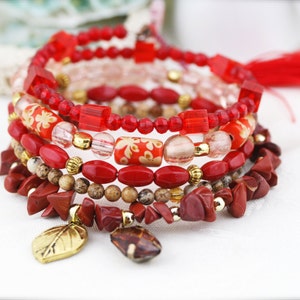 Puede incluir: Un conjunto de seis pulseras rojas y doradas con un dije de hoja. Las pulseras están hechas de varios materiales, incluyendo cuentas, piedras y metal.