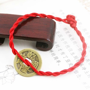 May include: A red braided bracelet with a bronze coin charm. The coin has Chinese characters and a square hole in the center. The bracelet is knotted at the end.